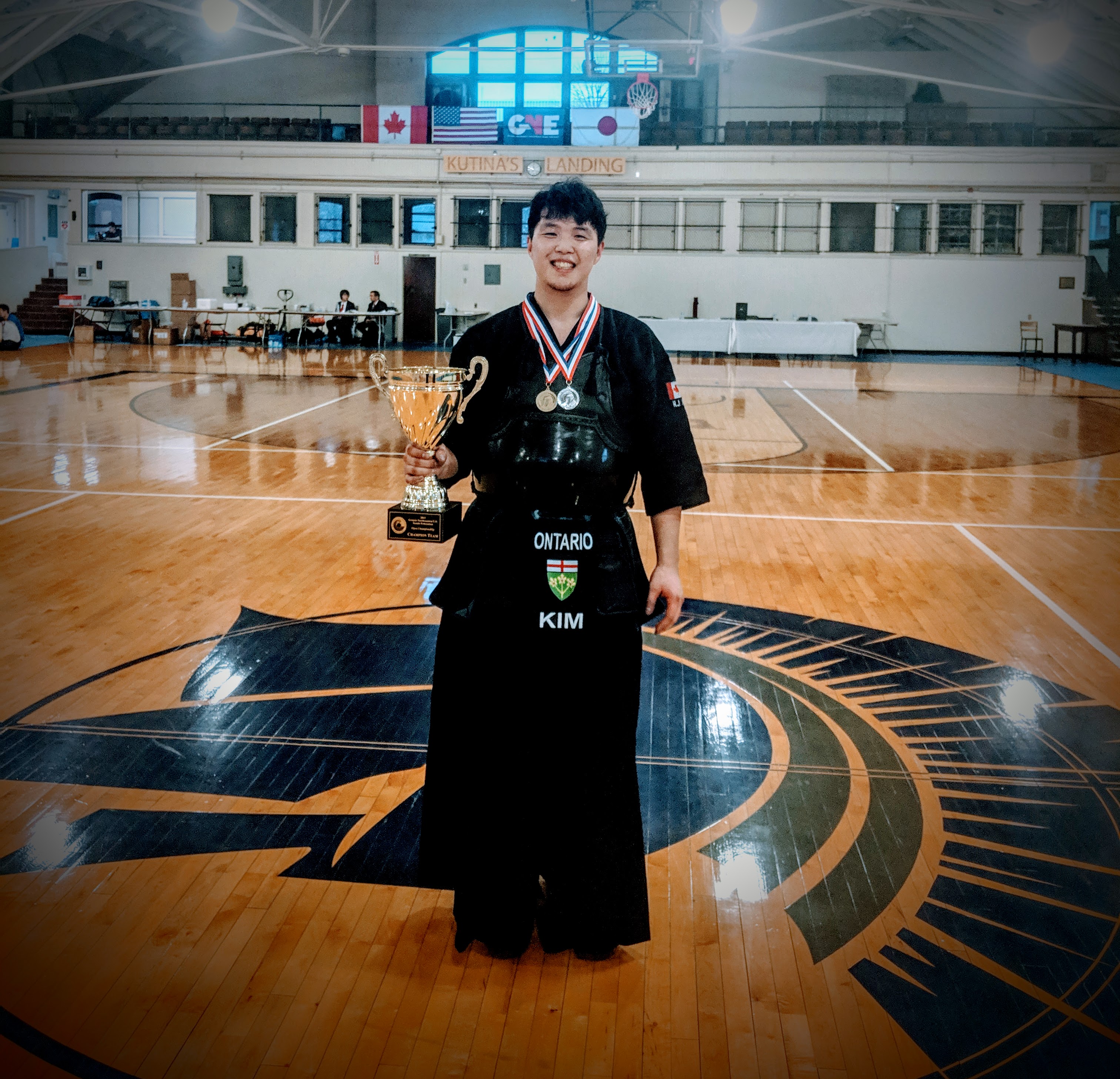 Instructor, Bill Kim, standing in a gymnasium wearing a kendo uniform and bogu (armour) and holding a trophy